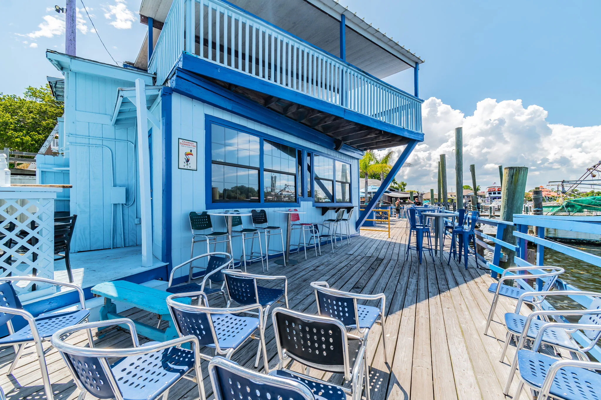 The outdoor wooden deck of the restaurant, featuring blue and black chairs and a view up to the second-story balcony.